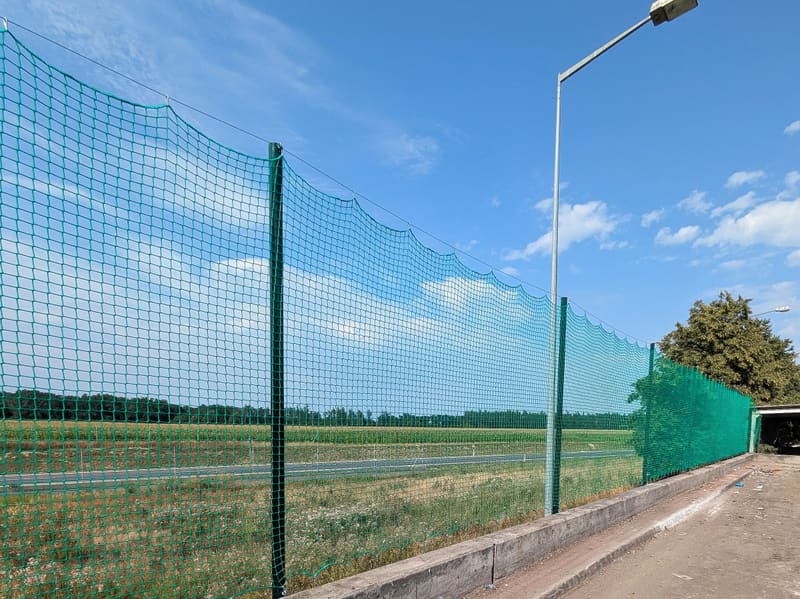 Wysokie ogrodzenie z zieloną siatką ochronną na stalowych słupach wzdłuż drogi dojazdowej